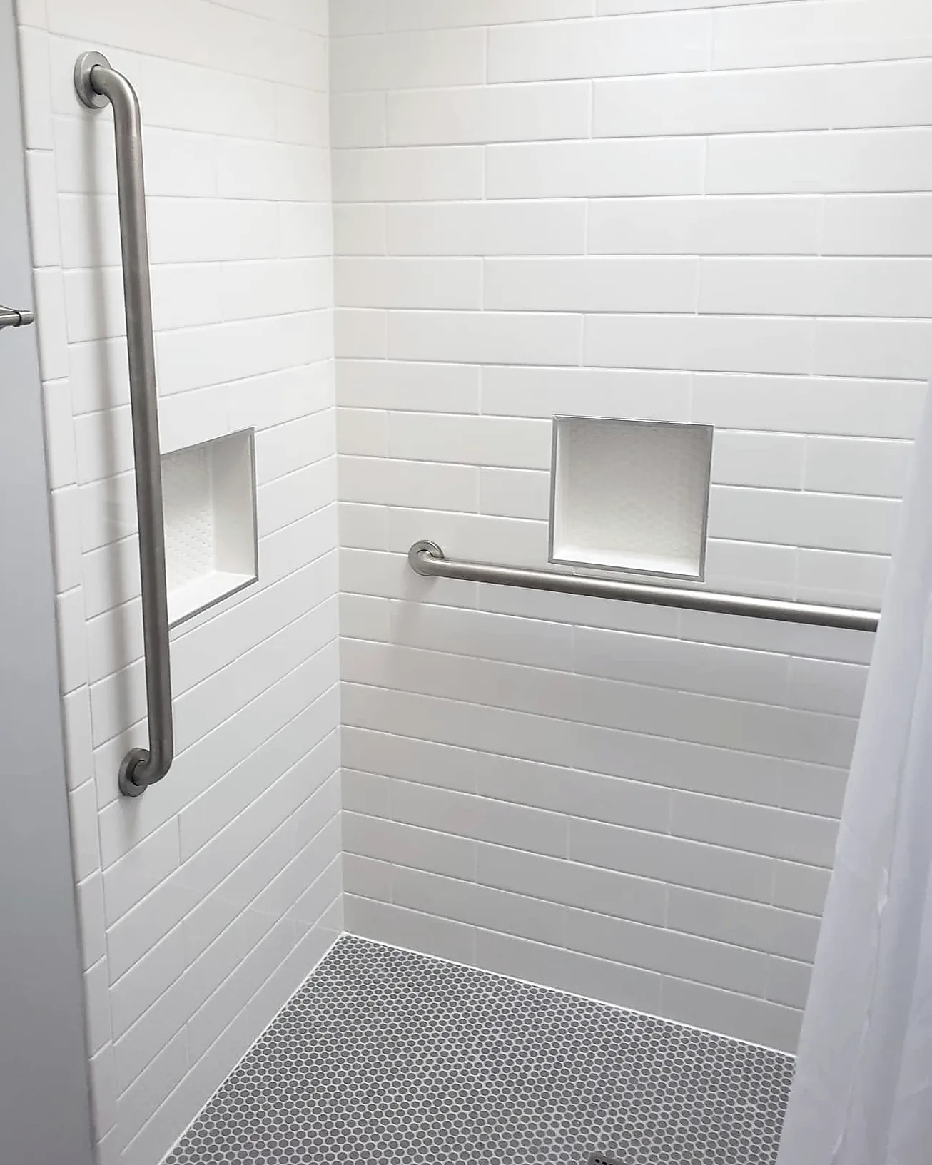 Inside a home’s accessible shower: white tile, a vertical and a horizontal grab bar, two tiled niches and a grey penny-tile floor.