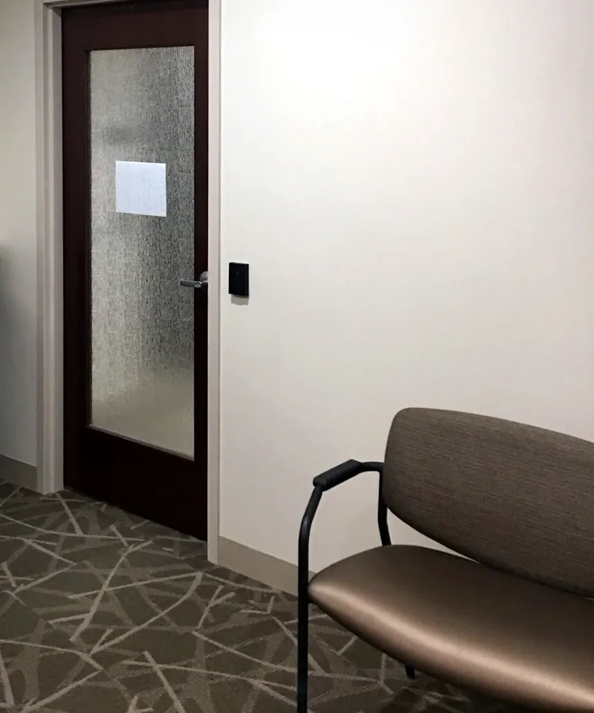 The door Patton moved forward: a dark door with a frosted glass panel and a card reader on the white wall, with a waiting-room chair beside it.