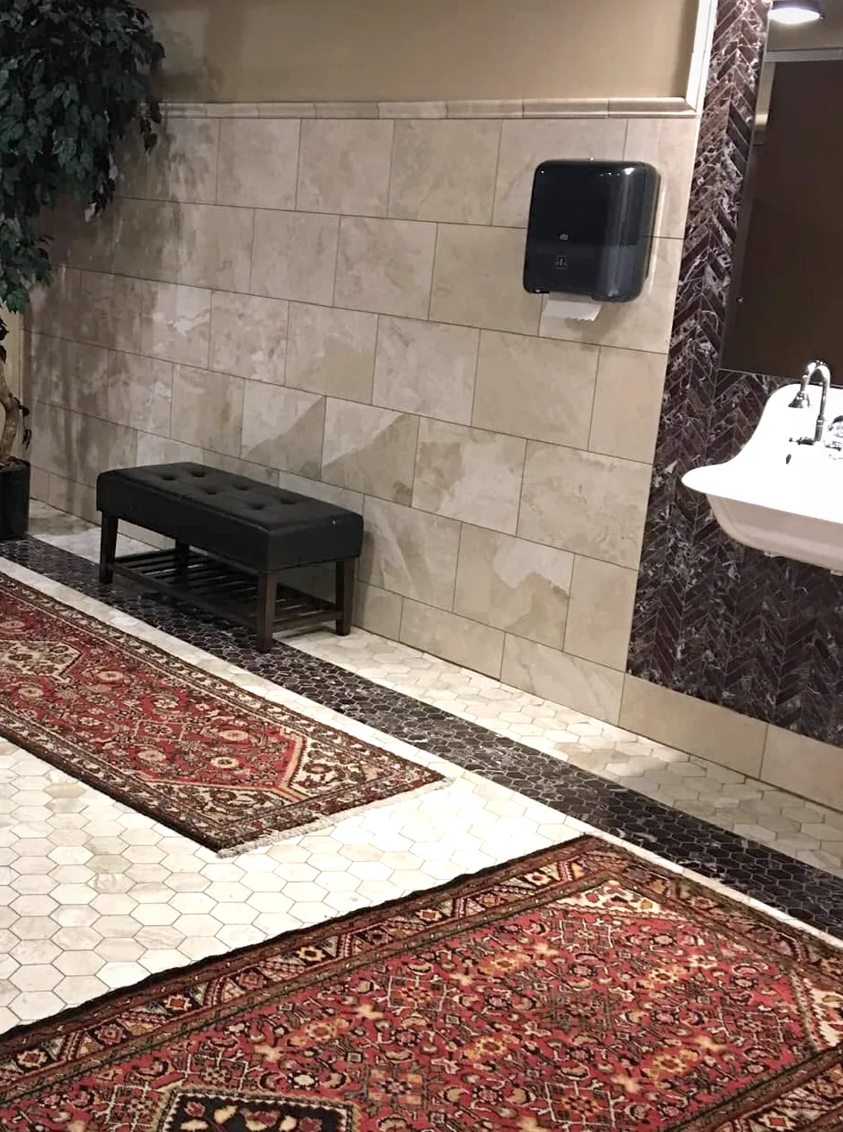 The same restroom: a bench under the beige wall tile, the hex mosaic floor and its dark border, and a runner rug.