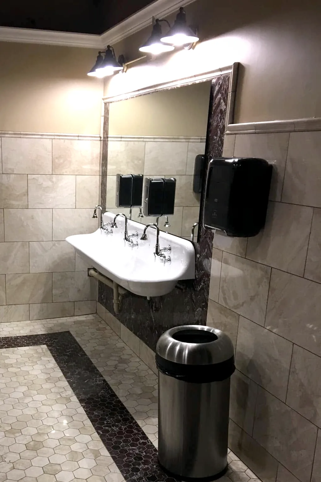 The finished restroom: three lit sconces over a wide mirror, a white trough sink with three faucets, beige wall tile, and the hex mosaic floor with its dark border.