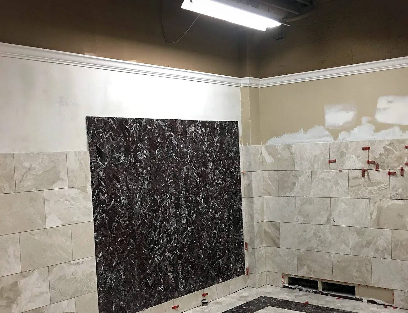 mid-job: large wall tile half laid beside a dark stone panel in a commercial restroom, the spacers still in the joints.